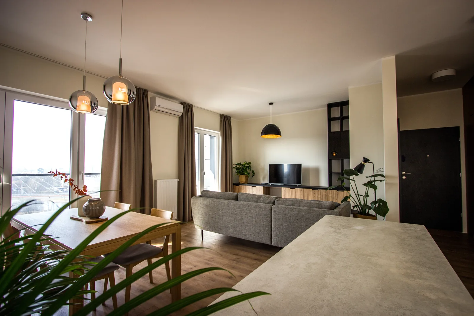 Open-plan dining and living panorama with oak table, chrome globe pendants, mountain art prints, and grey sofa with gold-interior dome pendant beyond