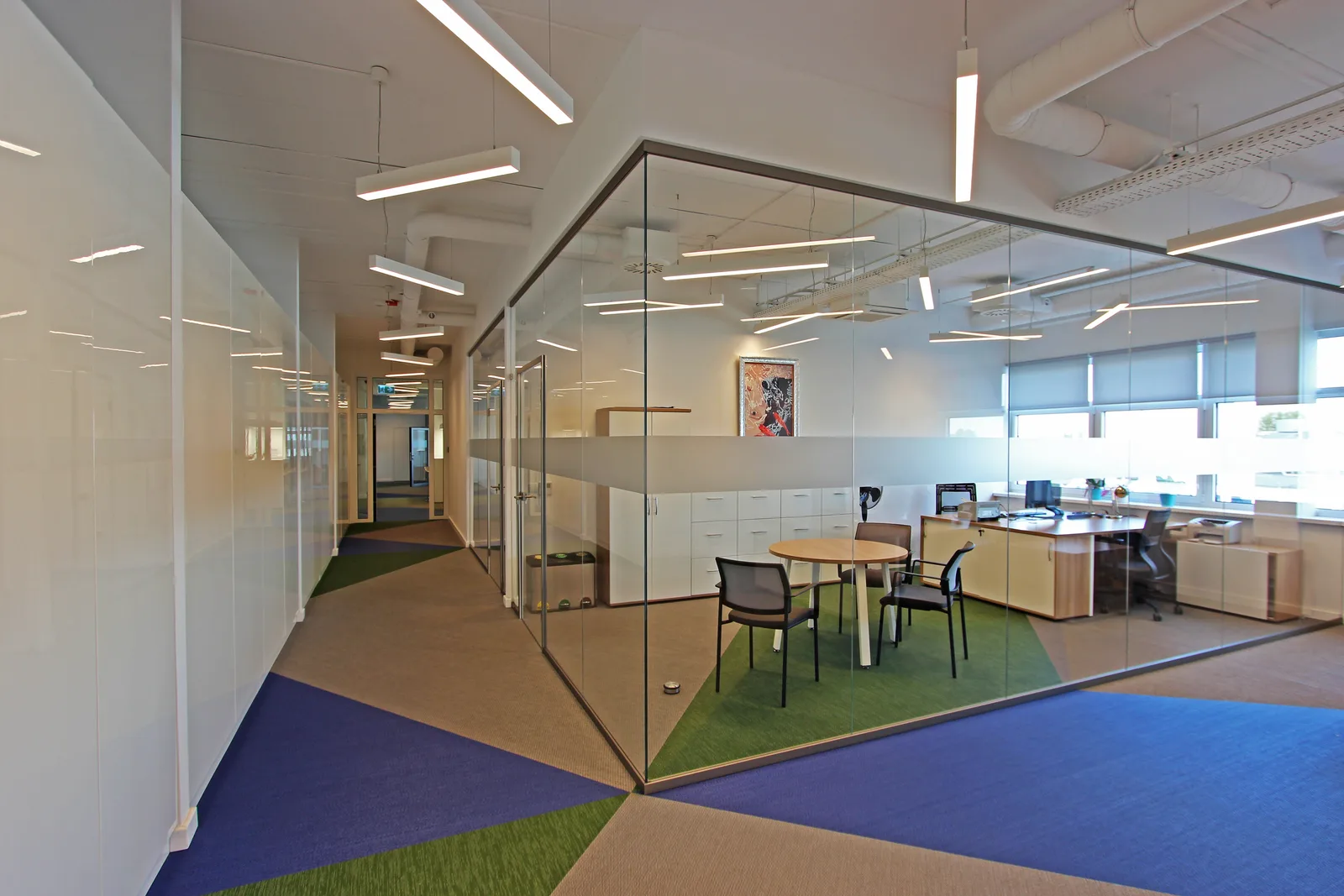 Corridor with glass-walled office view showing frosted band, three-colour woven vinyl floor pattern wrapping through corridor into office, and meeting corner
