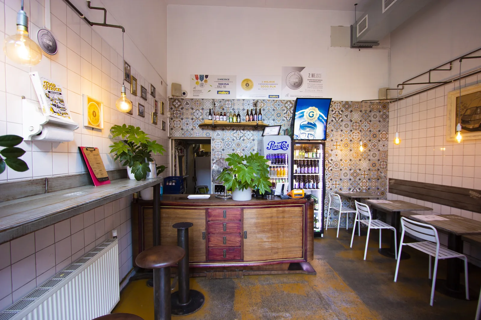 Ground floor deli and bar interior with decorative patchwork cement tiles as wall feature, vintage wooden counter with plants, industrial pendant lights
