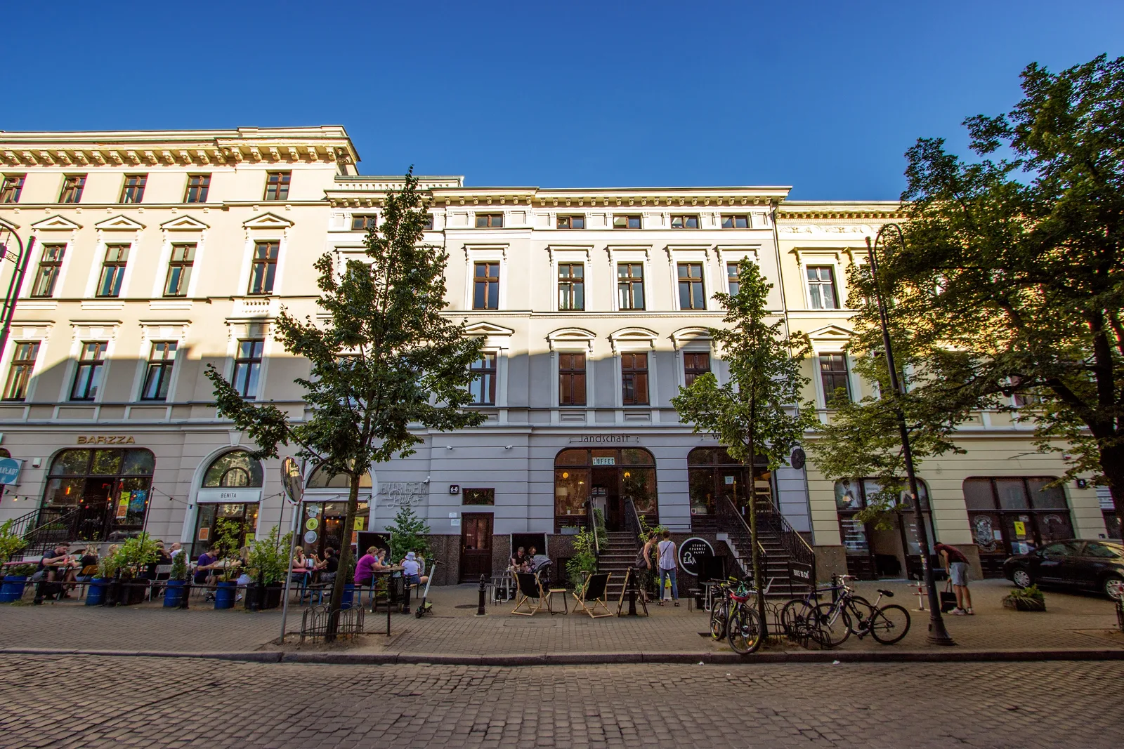 Wide summer street panorama showing full facade with commercial storefronts, trees, bicycles, cobblestone