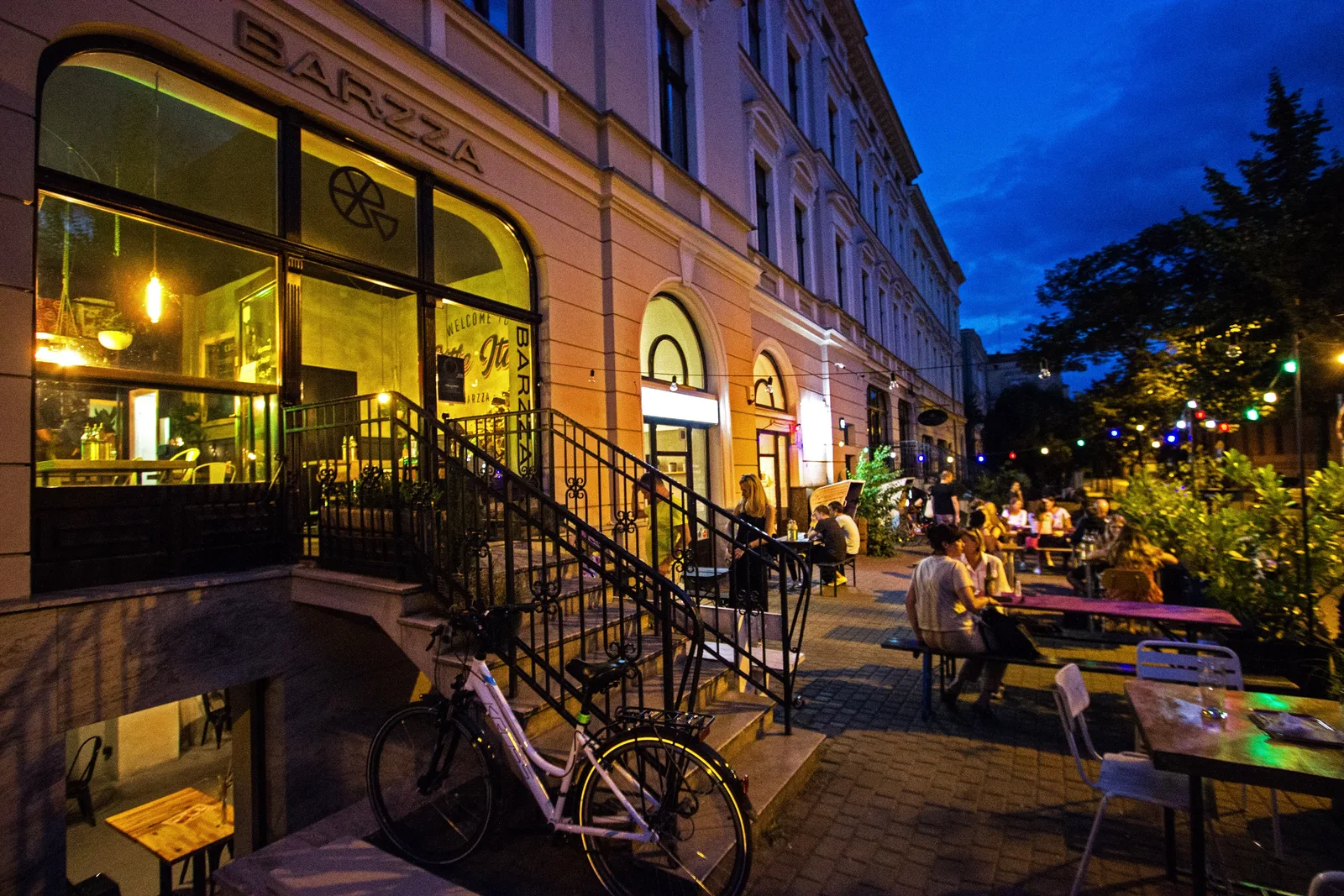 Evening dusk street scene with BARZZA cafe illuminated, warm lighting, iron staircase railings, outdoor dining tables, deep blue sky