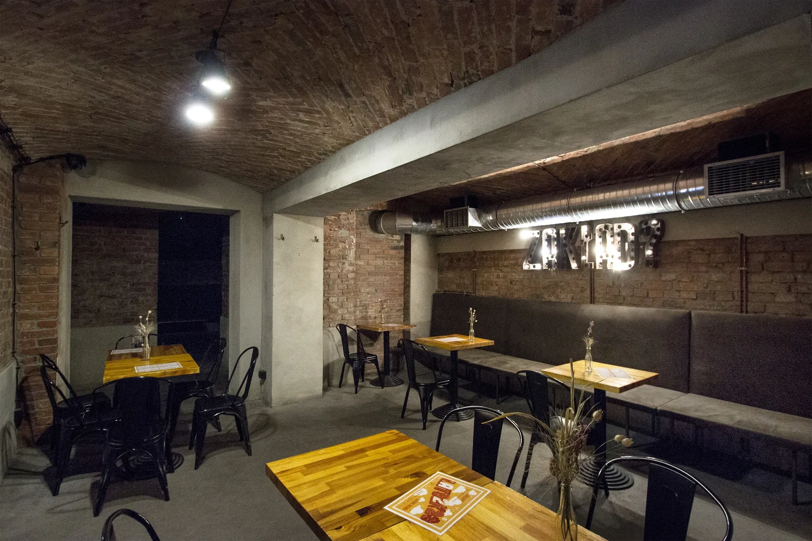 Basement vault restaurant with exposed concrete and brick ceiling, industrial pendant lights, yellow wooden tables, banquette seating, GYMSZT wall text