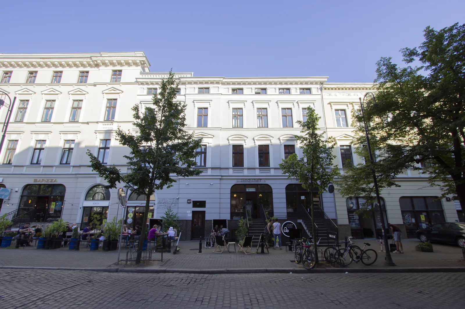 Wide summer street panorama showing multiple restored neoclassical buildings, pedestrians, outdoor cafe seating, bicycles on cobblestone pavement