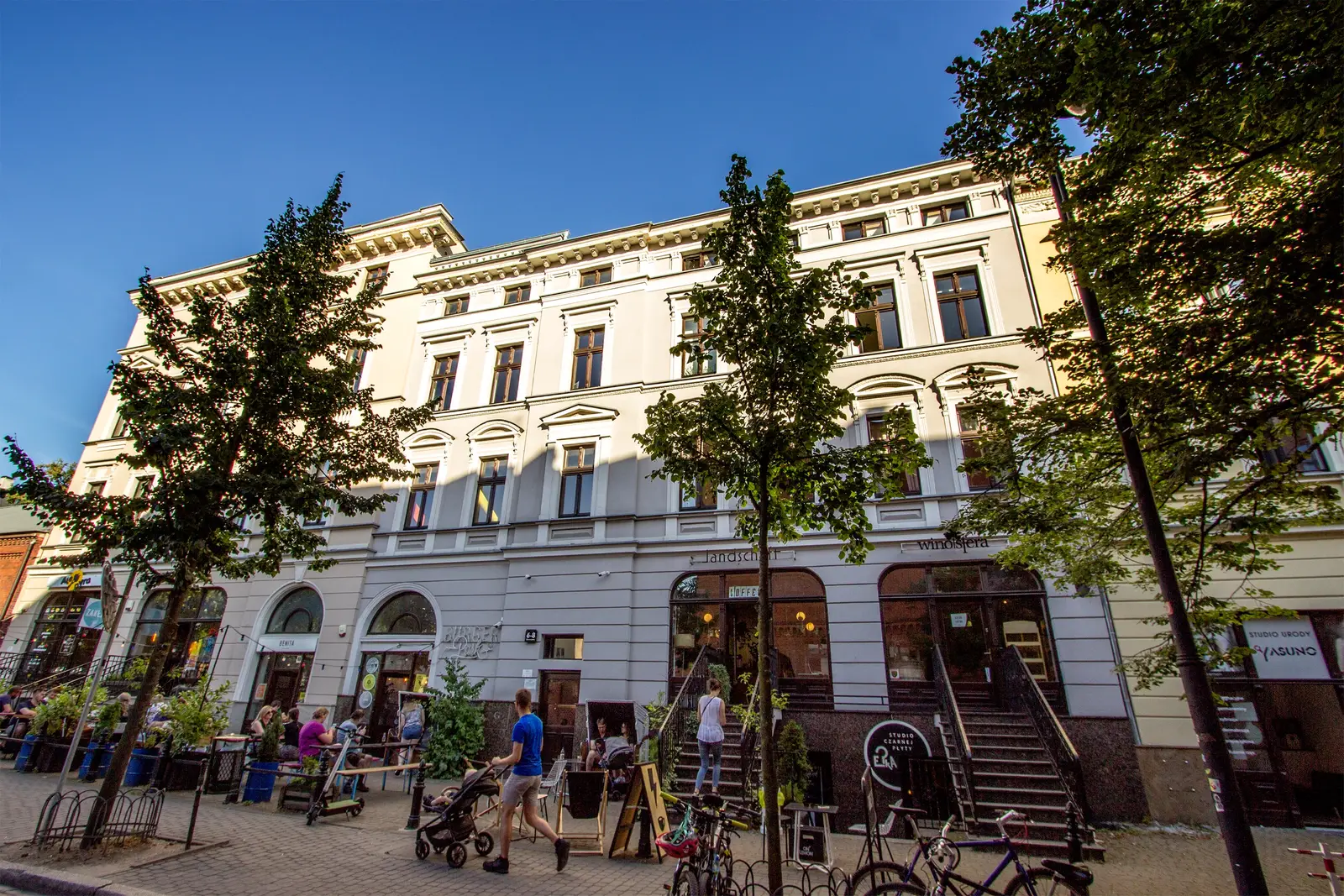 Angled close-up of main building facade from low viewpoint, full-height neoclassical windows, commercial storefronts at ground level with Landschaft signage