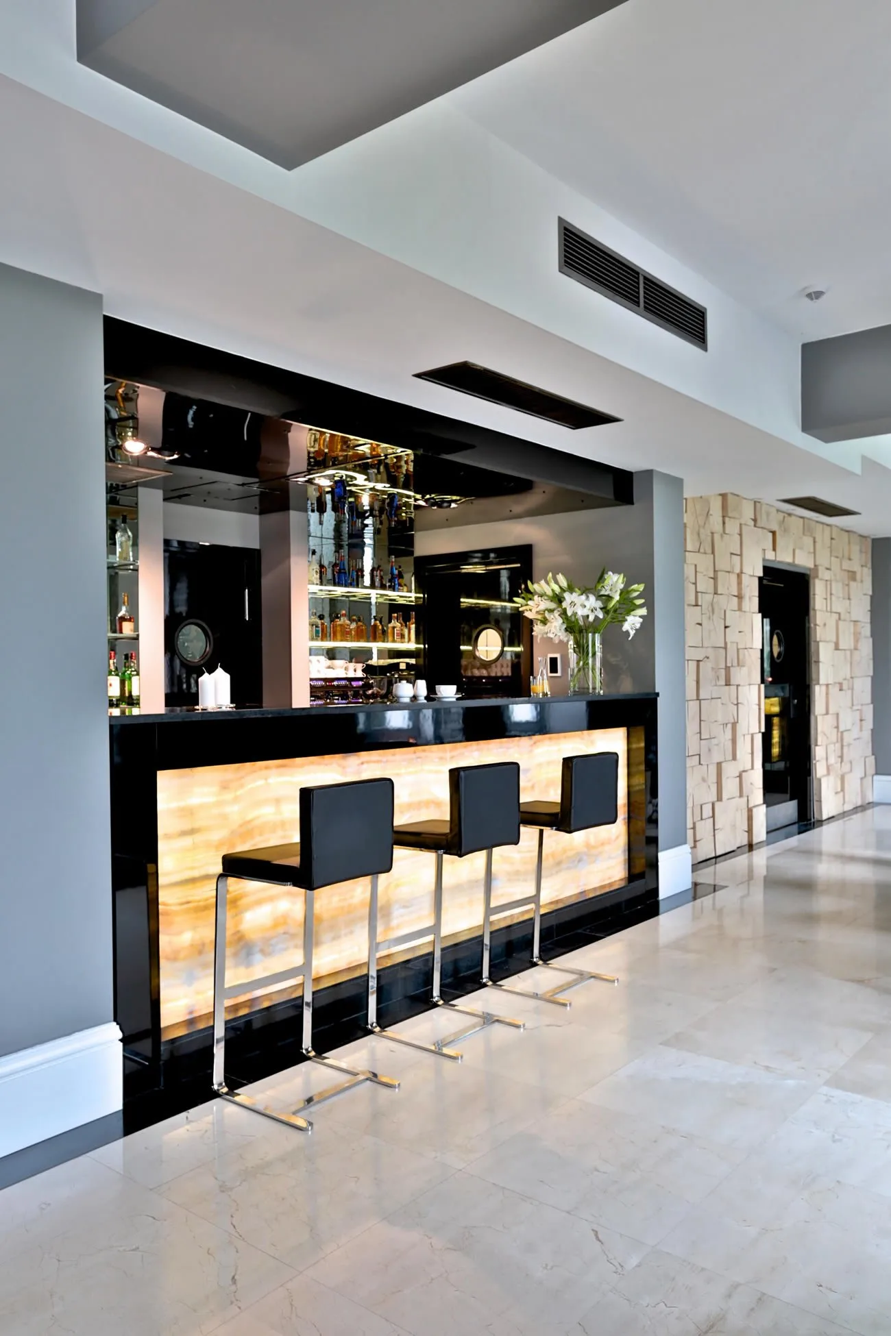 Backlit onyx bar element glowing warm amber alongside natural stone wall, with Panton chairs and a mirrored wall behind