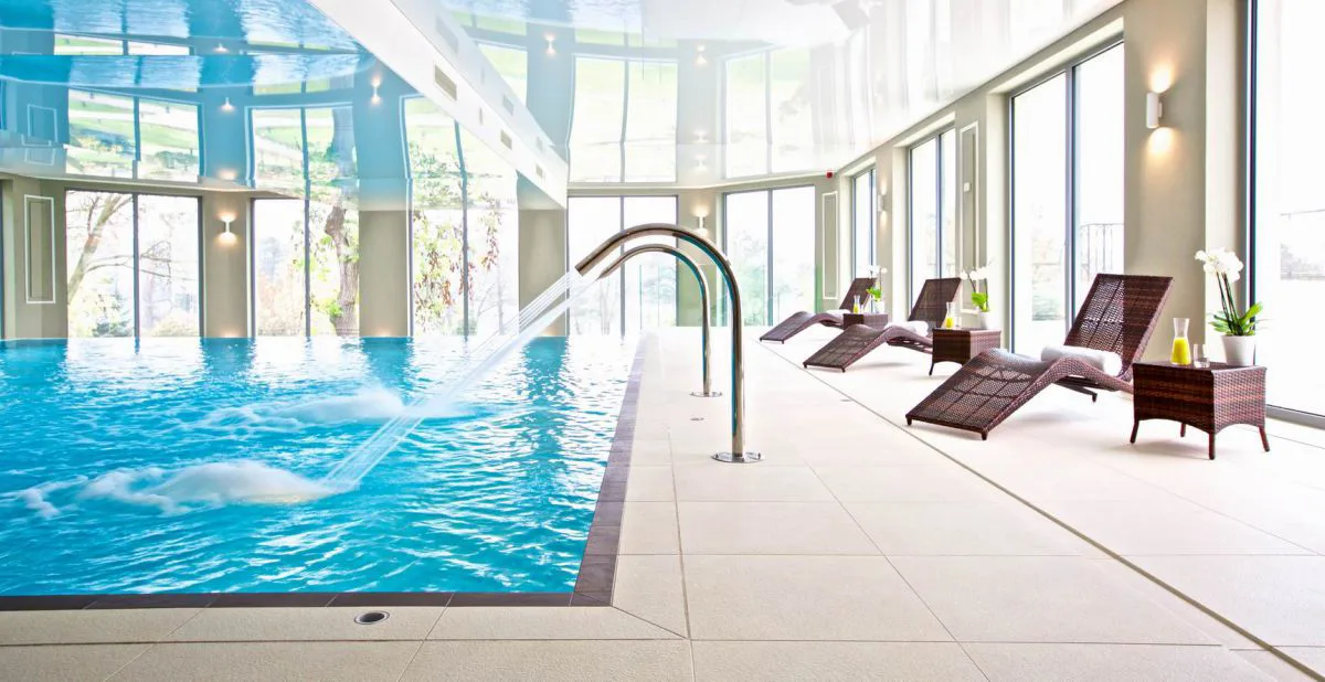Infinity pool side view with stretch ceiling, loungers, water features, and landscape visible through glazing