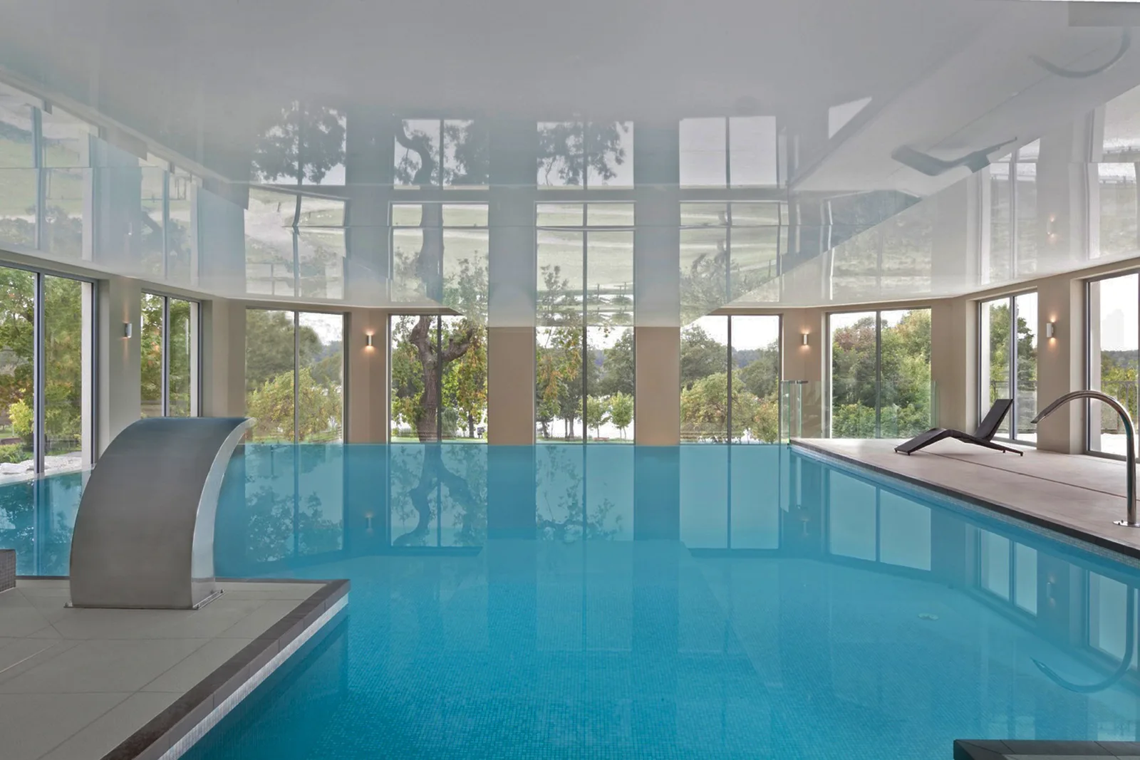 Infinity pool interior with stretch ceiling, steel waterfall cascade, panoramic windows, and poolside loungers