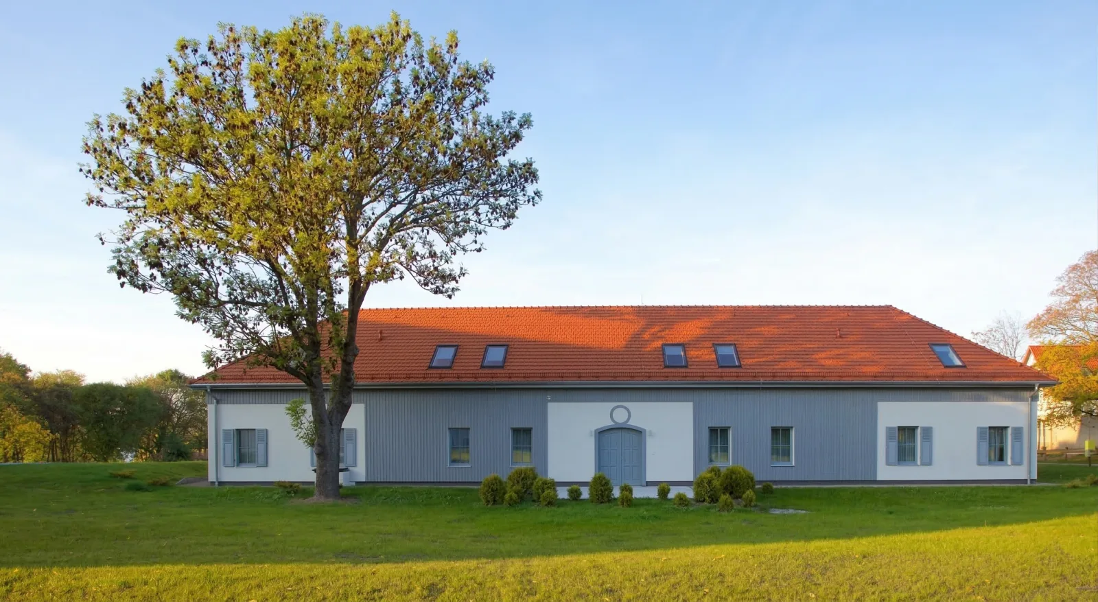Annexe and stables building exterior in summer with white walls, red tile roof, and single tree against blue sky