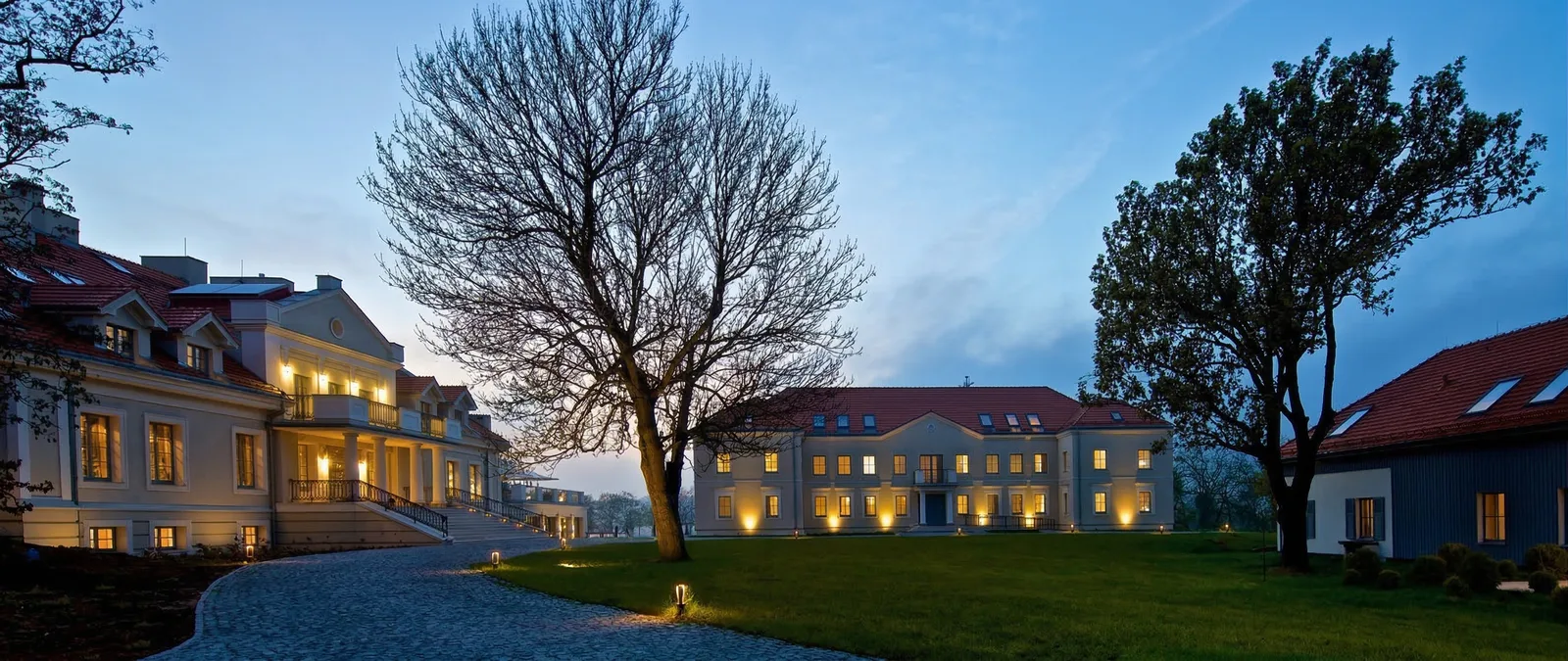 Twilight estate panorama with three buildings illuminated at dusk, pathway lights tracing routes through the landscaped park