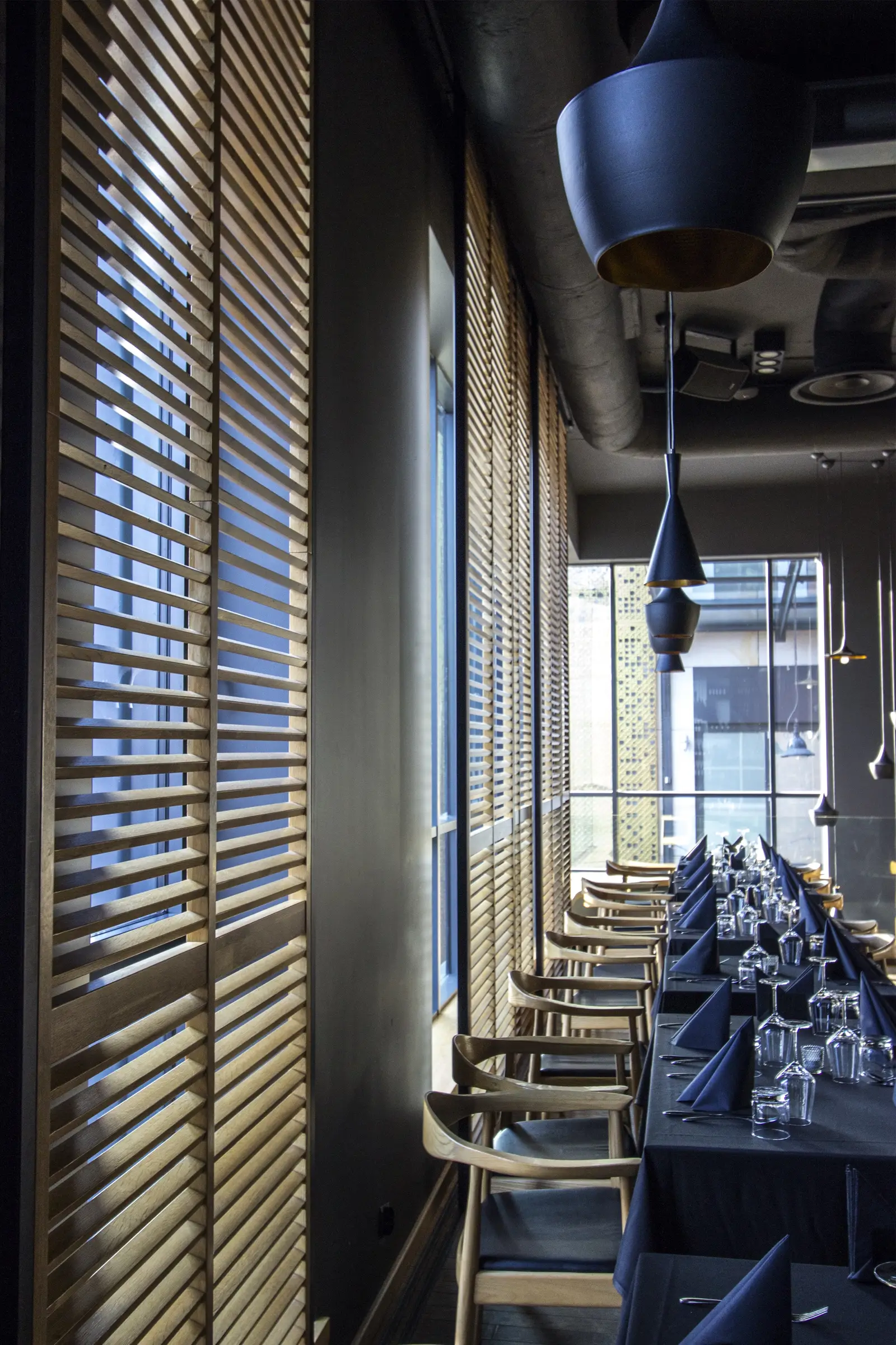 Detail: oak venetian blinds and Tom Dixon pendants close-up with warm light through slats and dining tables visible