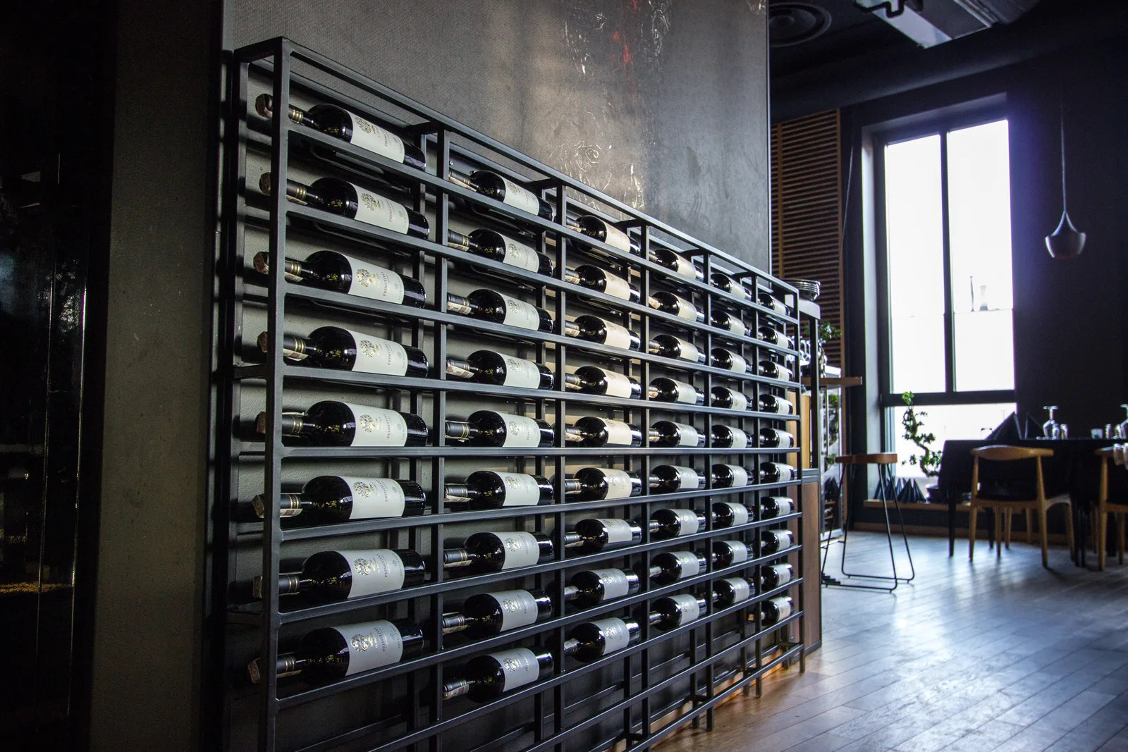 Detail: black metal wine rack with industrial horizontal bottle storage, wall-mounted, and ambient lighting from dining area