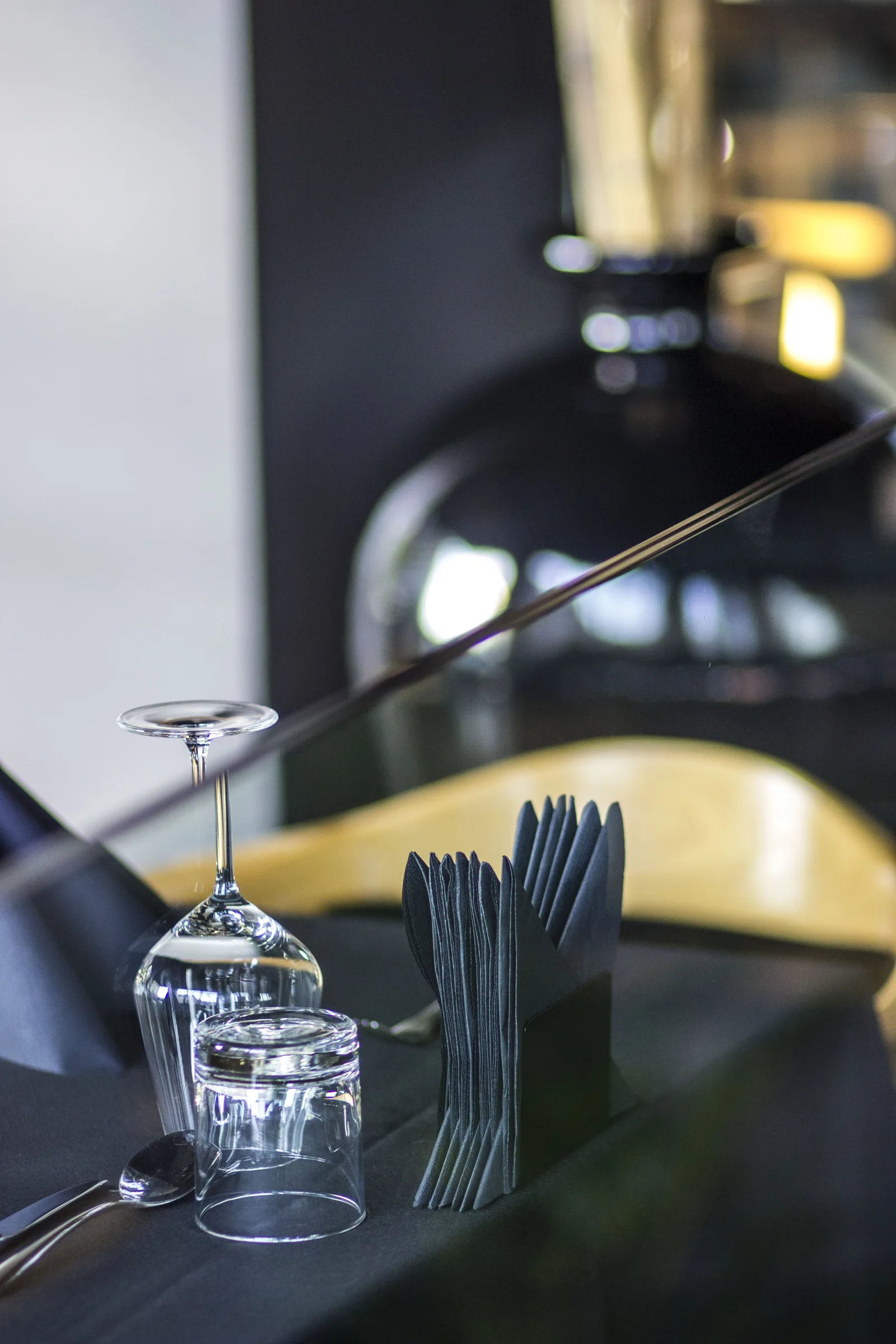 Detail: table setting close-up with inverted wine glass, black napkin fan in holder, dark tablecloth, and Tom Dixon pendant bokeh background