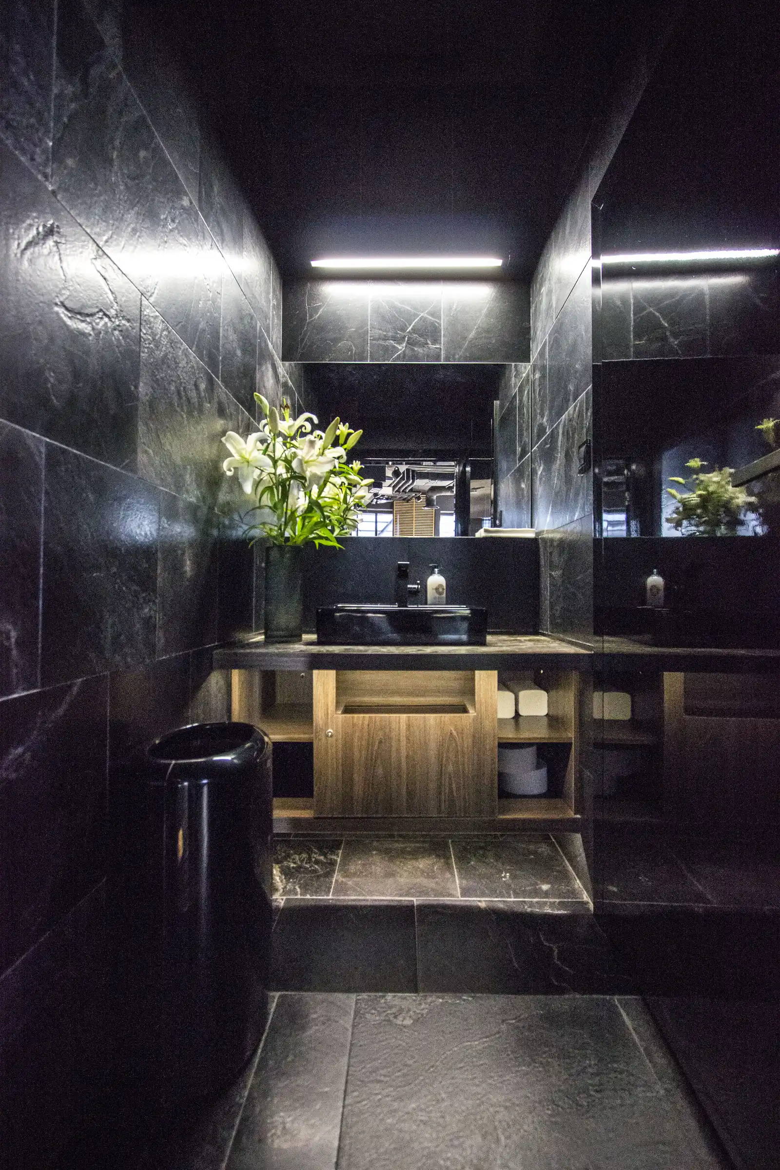 Interior: restroom with dark marble walls, white lily flowers, oak vanity with open shelving, and matte black basin