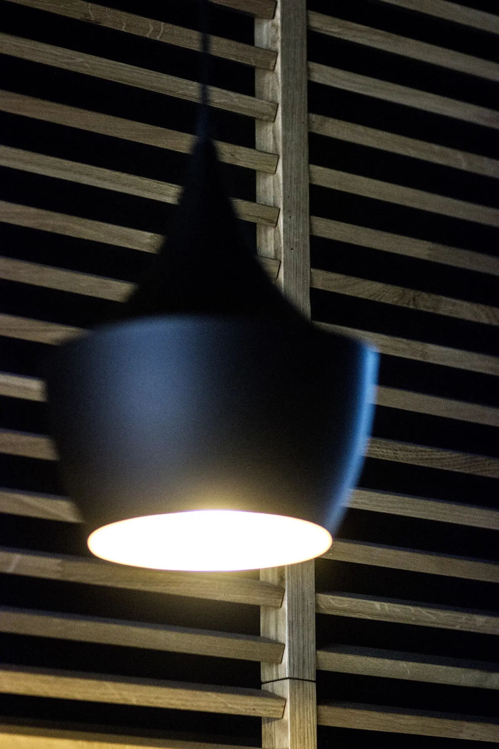 Detail: single Tom Dixon Beat pendant close-up illuminated against oak louver screen background
