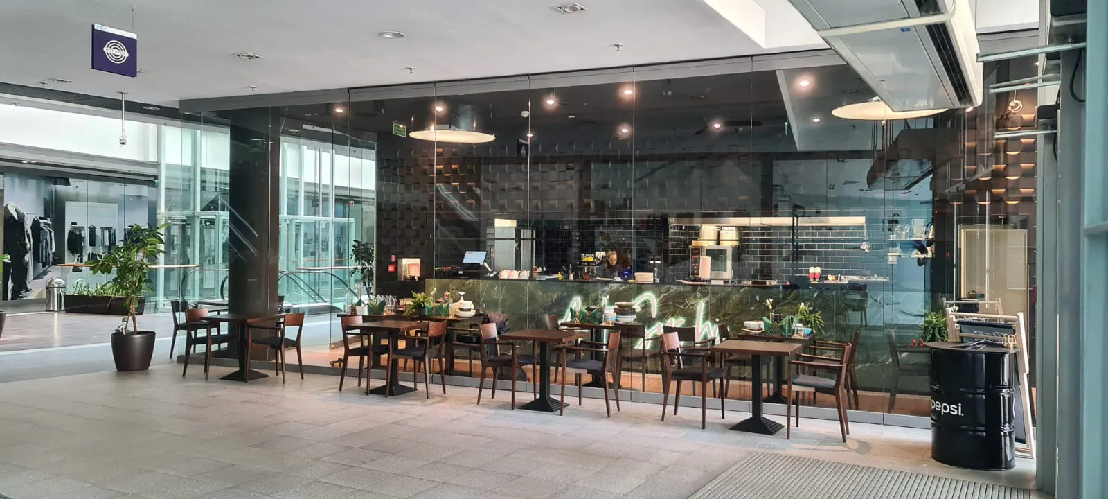 Exterior: through-glass photograph from wide left angle showing the complete built restaurant facade within Galeria Drukarnia mall — Verde Karzai bar, disc pendants, Fameg walnut chairs, and mall concourse context