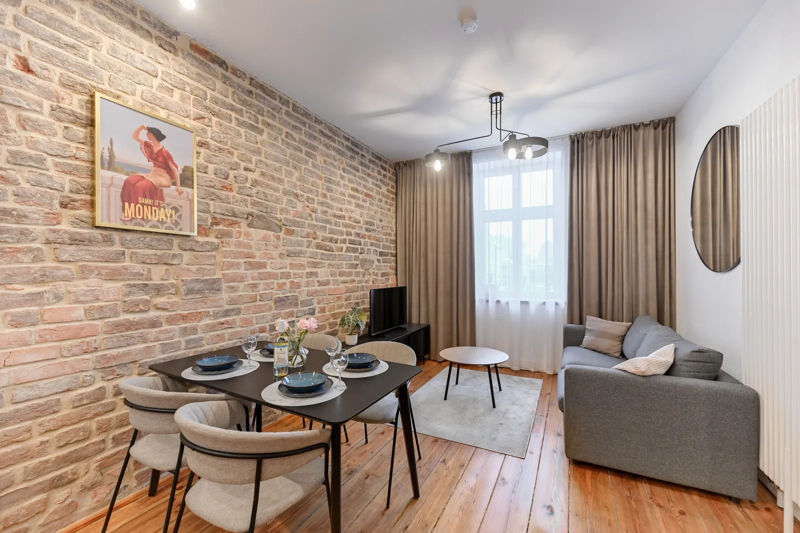 Interior: Apartment A signature shot with dining table, exposed brick accent wall, grey sofa, pendant lighting, and warm oak flooring throughout