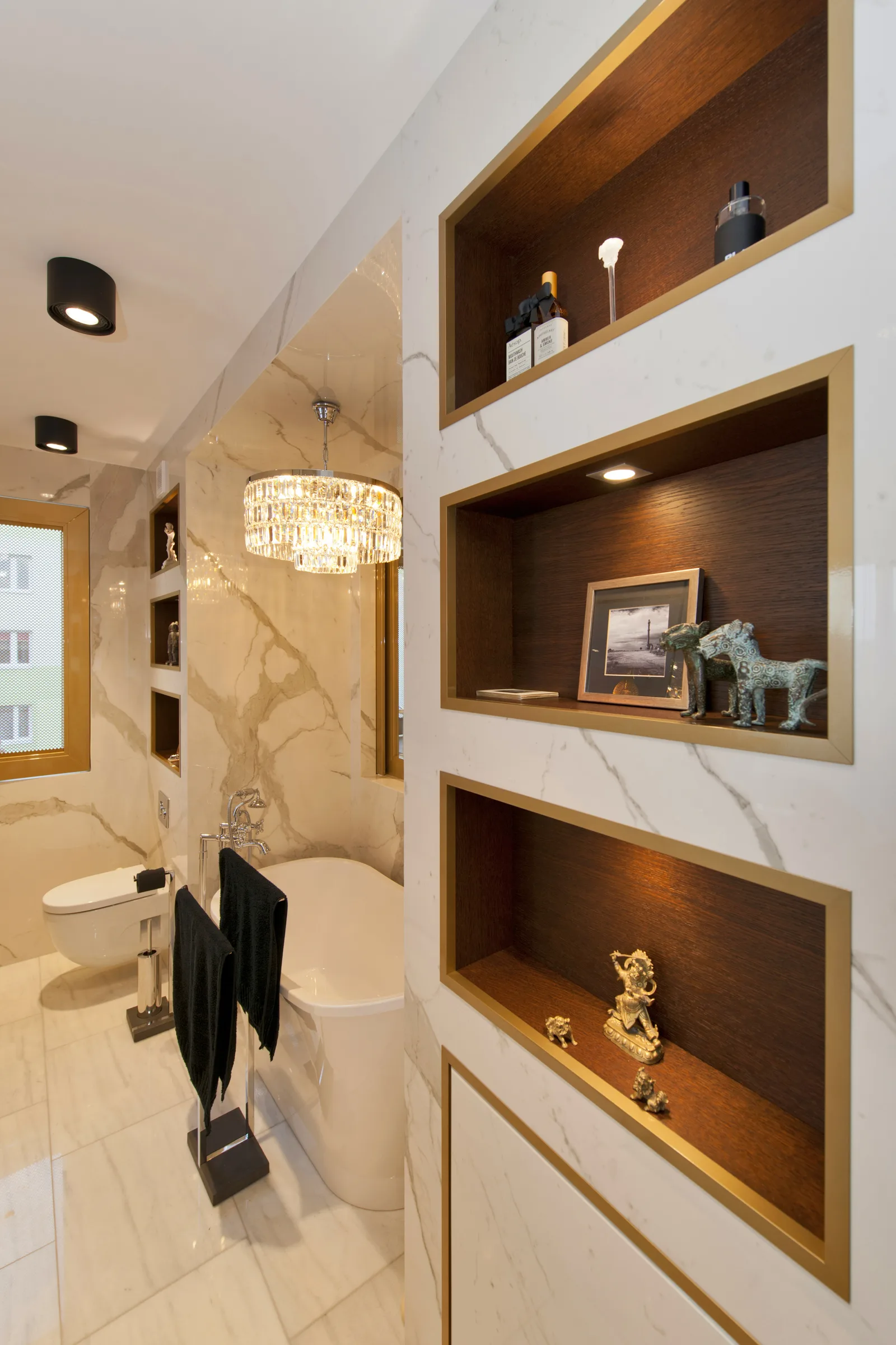 Bathroom shelving niches detail with marble wall, four recessed wood-backed shelves, crystal chandelier, freestanding tub, and decorative figurines