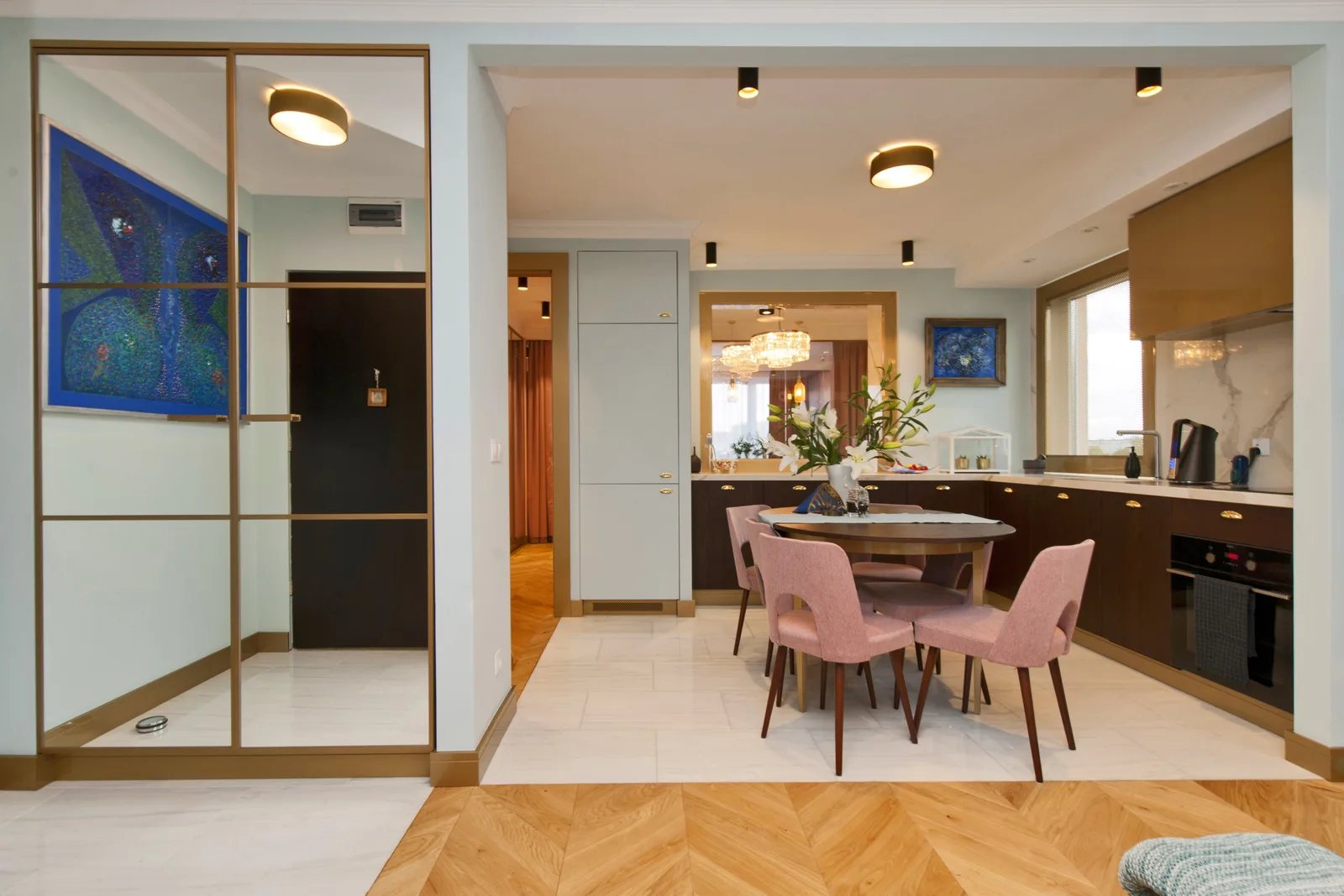 Kitchen-dining from entry with glass partition displaying blue abstract art, round dining table with pink chairs, dark wood kitchen with marble counter, and warm lighting