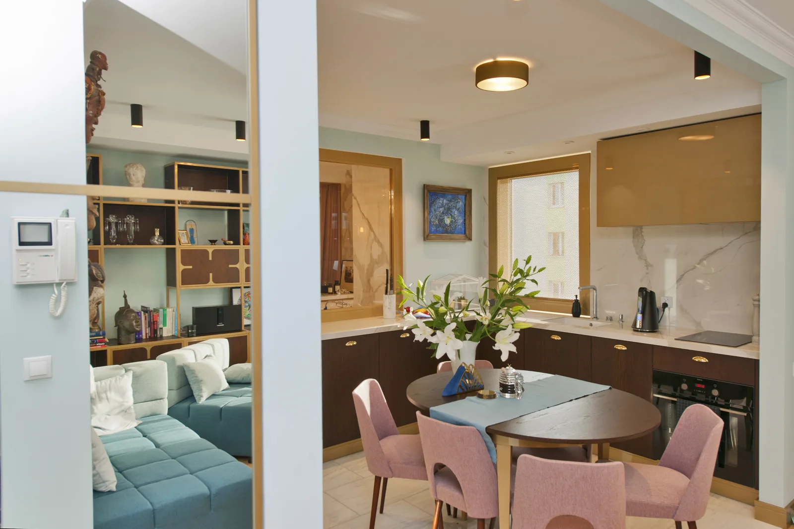 Living room through partition showing teal modular sofa, built-in bookshelf wall, glass partition framing, and pink dining chairs visible beyond