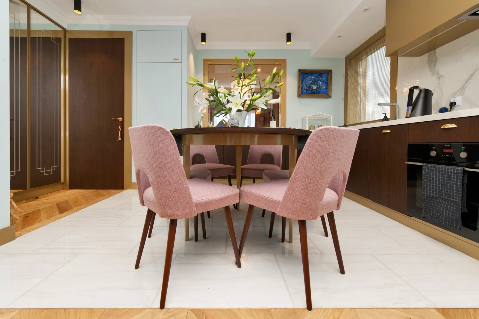 Dining close-up with pink velvet chairs around dark wood round table, white lilies centerpiece, dark wood kitchen cabinets with marble counter, and herringbone parquet