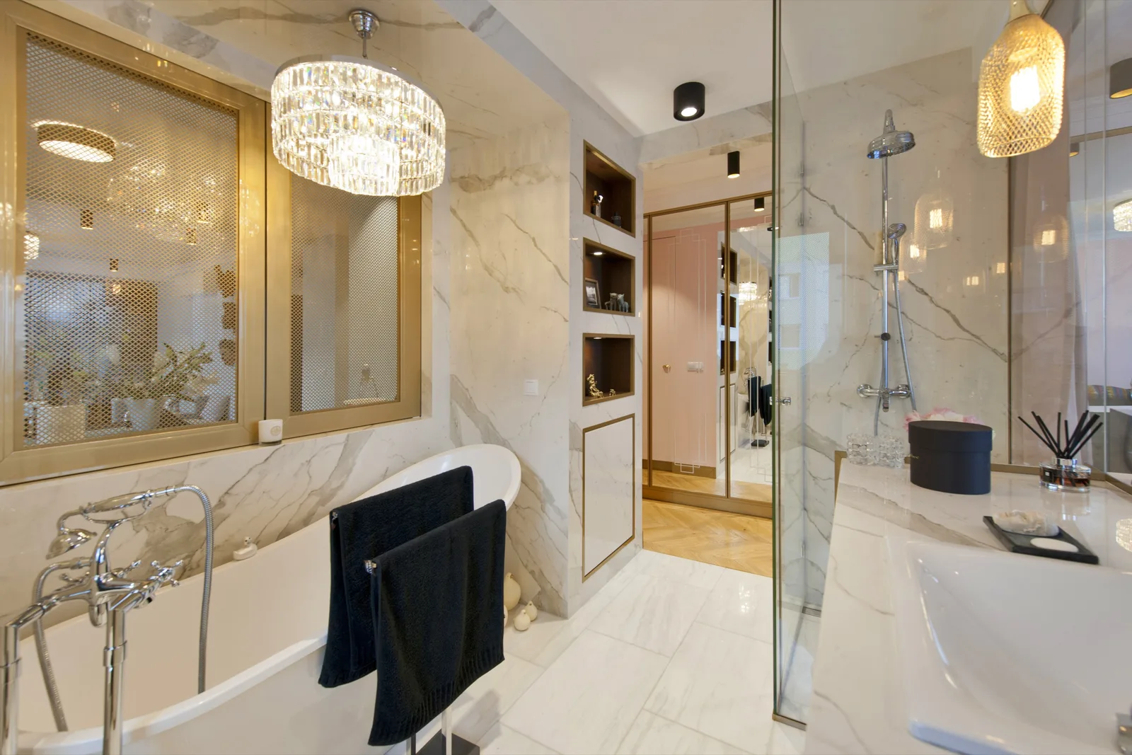 Luxury bathroom with freestanding bathtub, crystal chandelier, marble walls and floor, glass shower partition with brass framing, and recessed wood-backed shelving niches