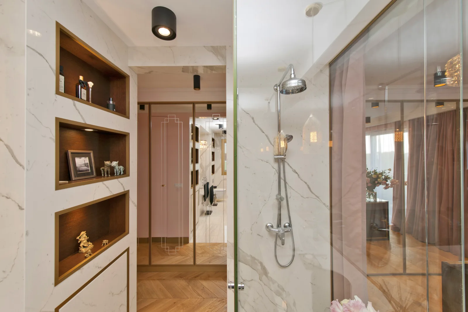 Bathroom corridor with marble wall, rain shower head through glass, recessed wooden shelving niches, and hallway to pink-toned room beyond