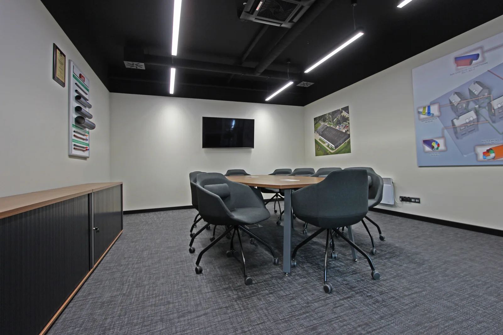 Interior: conference room wider angle showing walnut sideboard, product display poster, aerial factory photograph, and full boardroom table context