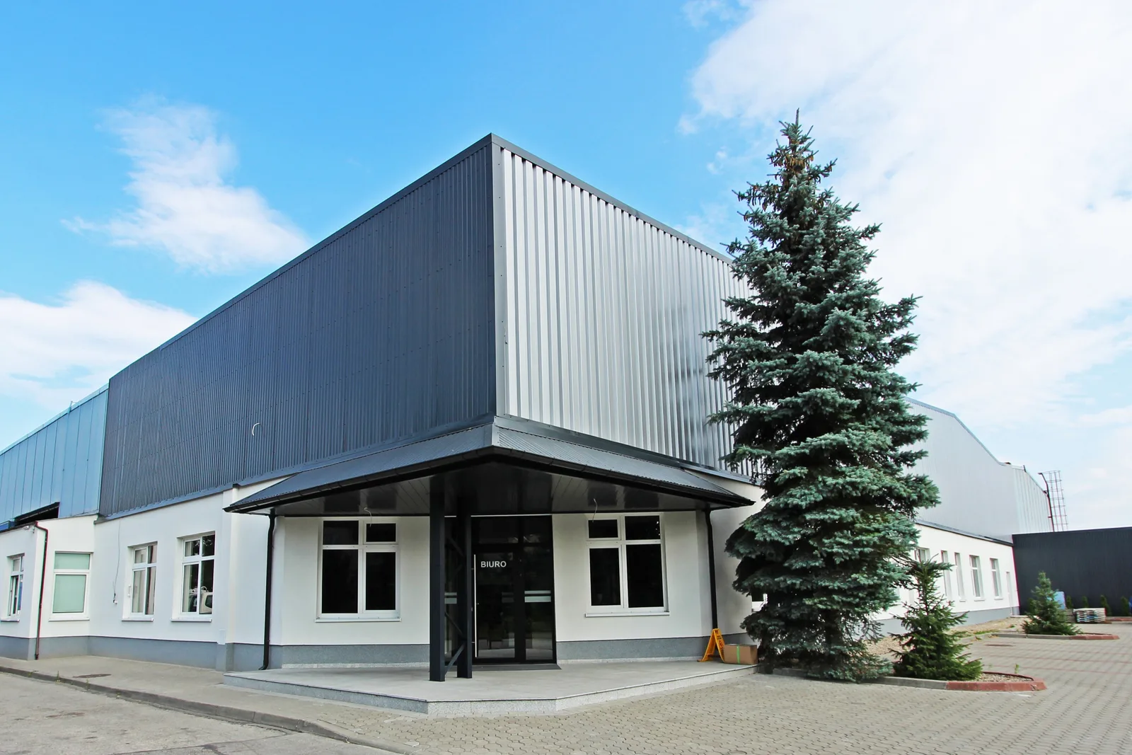 Exterior: front elevation of office building and production hall showing single-story white office block with BIURO canopy and silver-grey metal production hall