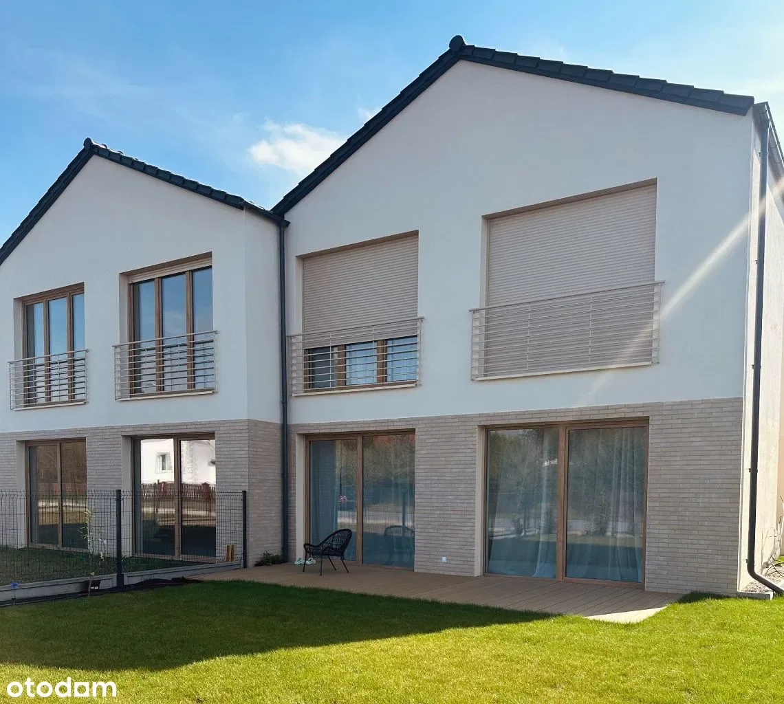 Rear garden realization photo showing floor-to-ceiling glazing, wooden deck terrace, green lawn, and clinker brick ground floor with white render upper