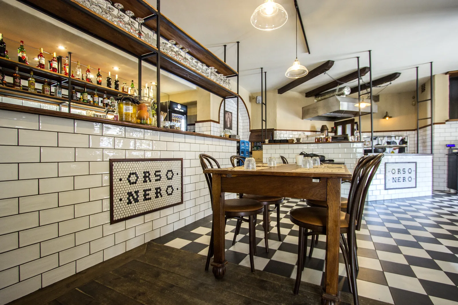 Interior bar zone with hexagonal tile mosaic spelling ORSO NERO on counter front, backlit bottle display, copper pendants, and reclaimed timber ceiling