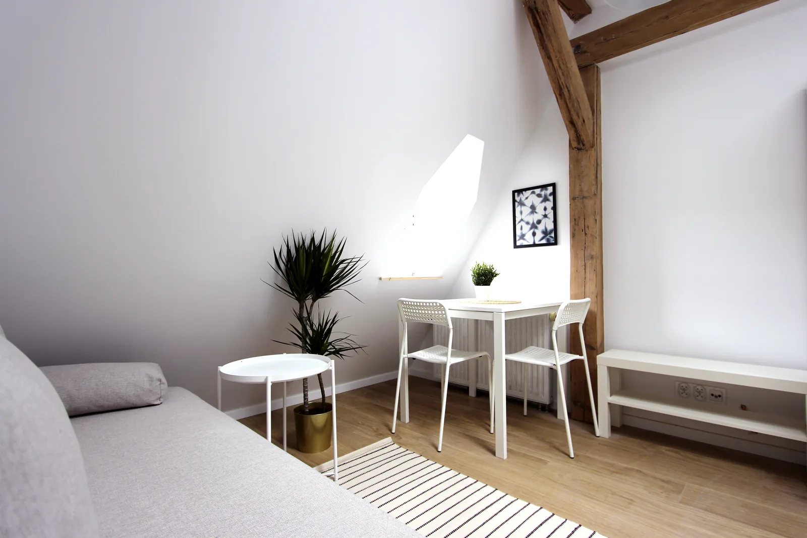 Detail: dining nook in dormer alcove with skylight above and exposed aged timber beam, showing how the attic gable geometry creates intimate spatial moments within the 25 square metre micro-apartment