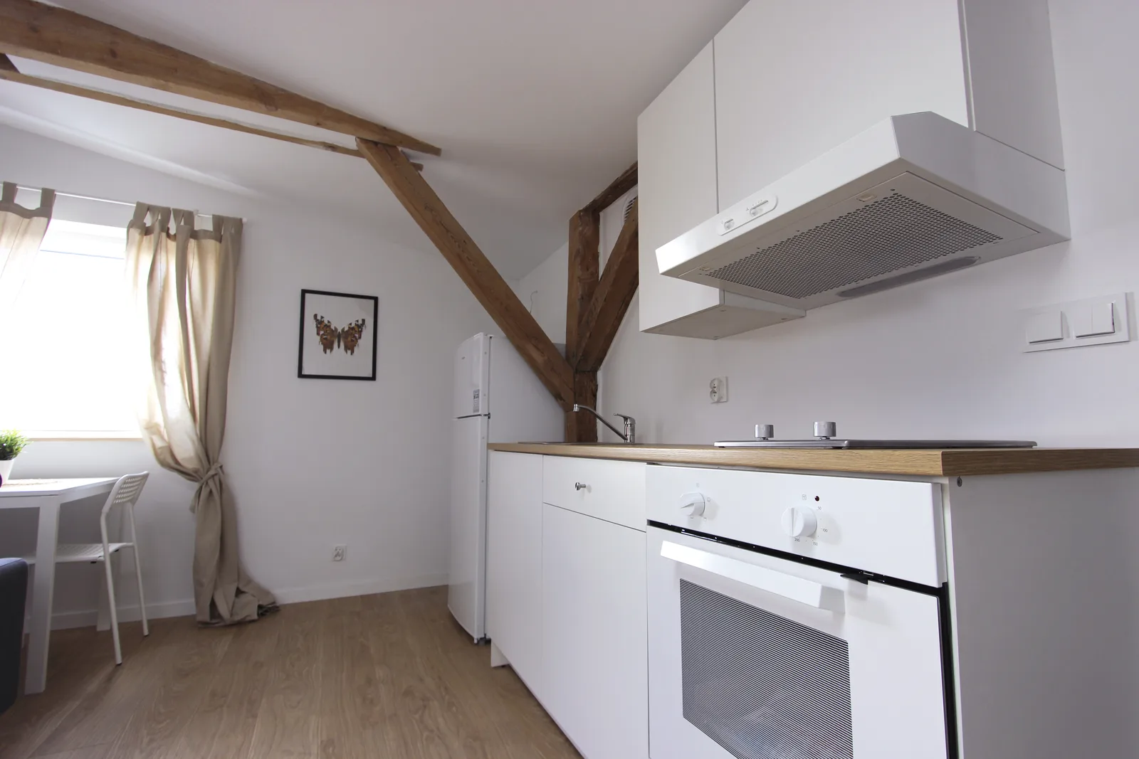 Detail: kitchen with exposed timber cross-brace beam overhead, white cabinetry, light oak laminate countertop, marble-effect splashback panel, and linen curtains at the dormer window