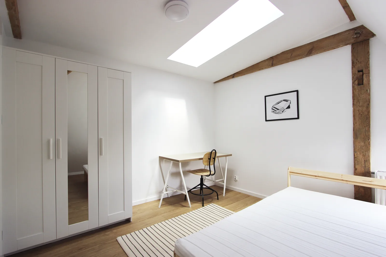 Interior: bedroom zone with wardrobe, desk, skylight directly above, exposed aged timber roof beams, and Ablux light oak laminate flooring throughout the Scandinavian-minimalist micro-apartment