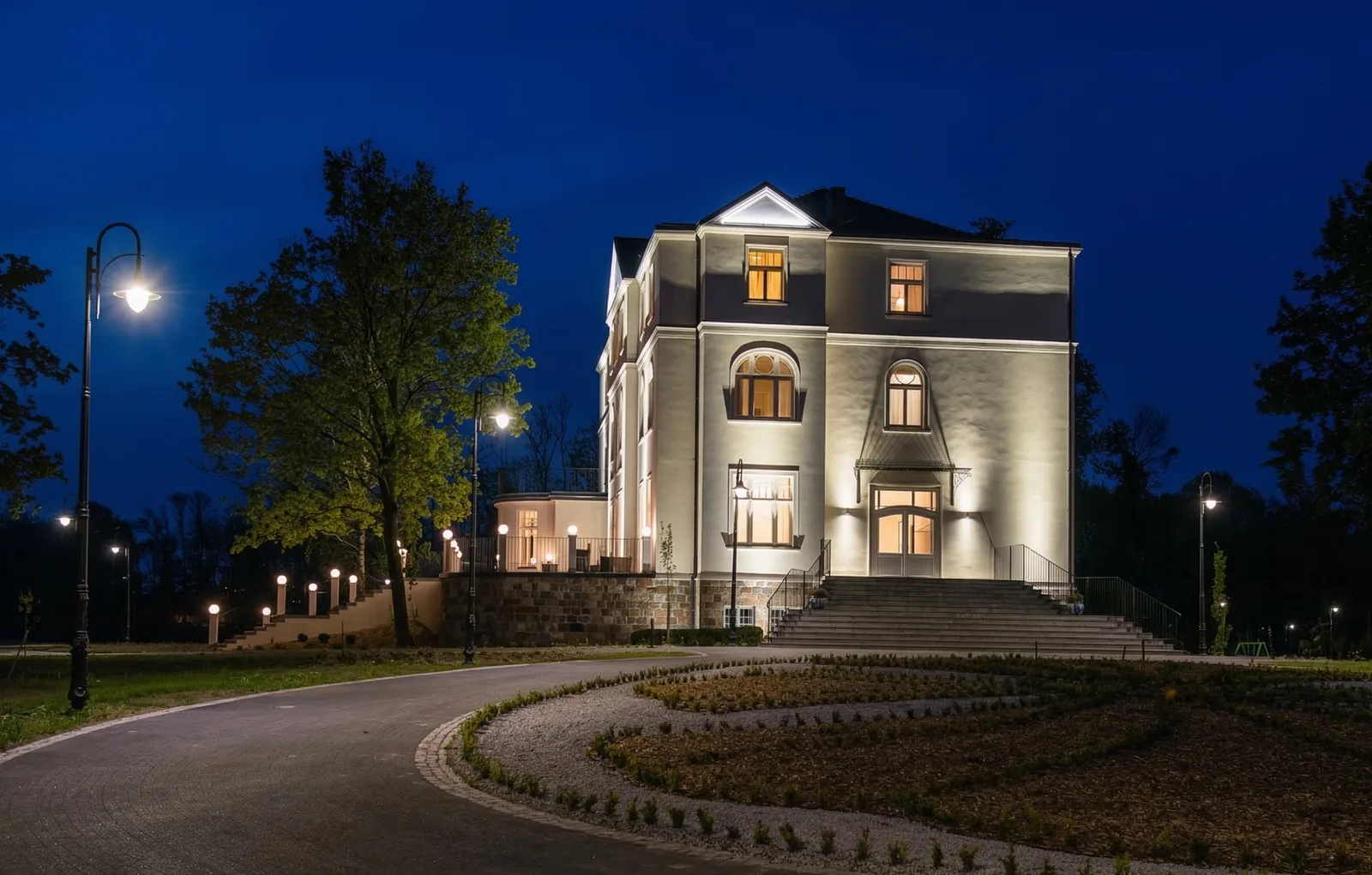 Night exterior of palace from driveway with round flower bed and warm street lamp lighting