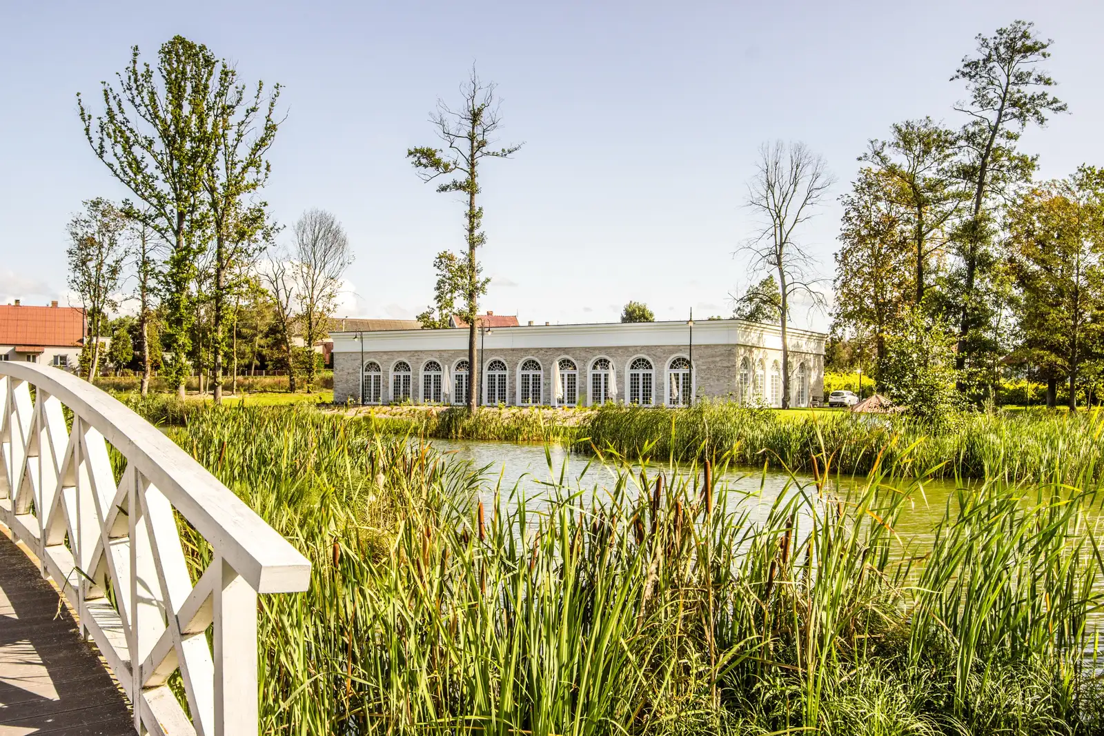 Orangerie from white wooden bridge with reeds, pond, and building in evening light
