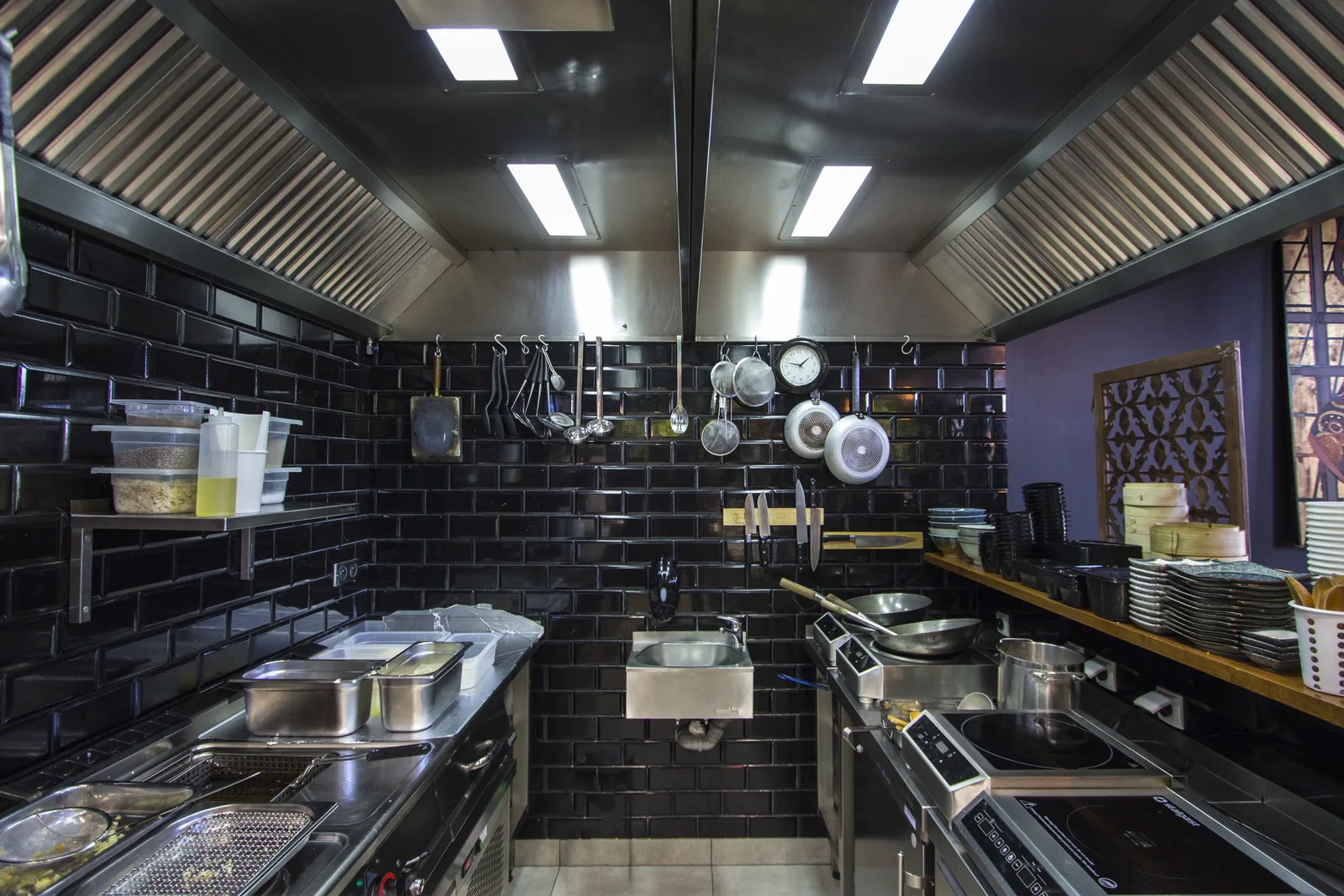 Interior: professional kitchen — stainless steel equipment, black subway tiles, induction cooktops, ducting