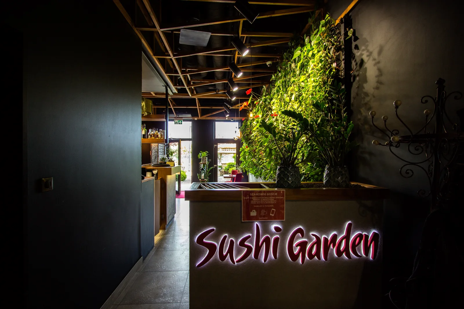 Interior: Sushi Garden reception — illuminated logo on counter, green wall behind, bar visible