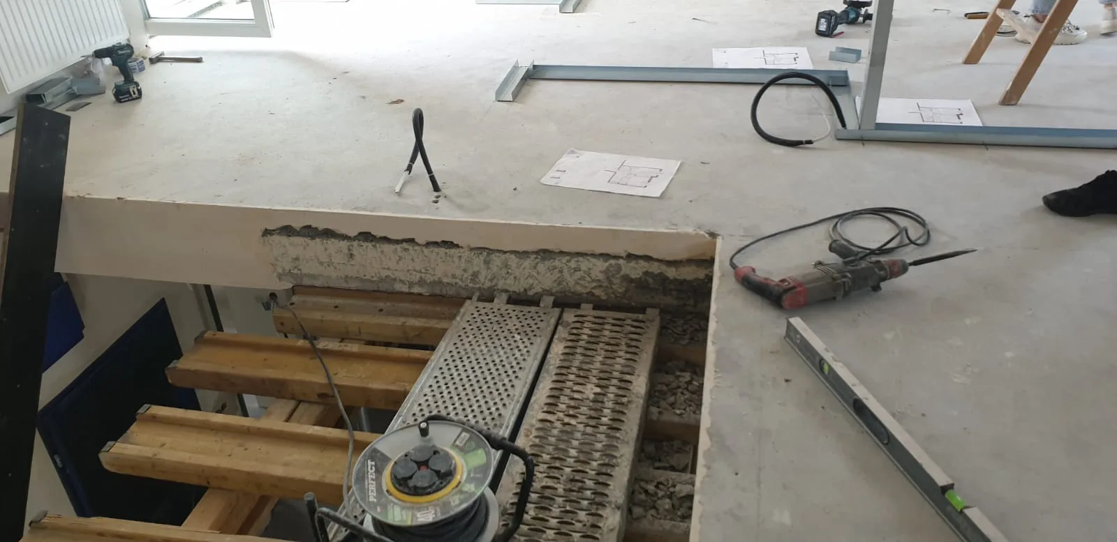 Slab opening from above showing cut concrete edge with exposed ribbed construction, power tools and cable reel on the floor during structural demolition
