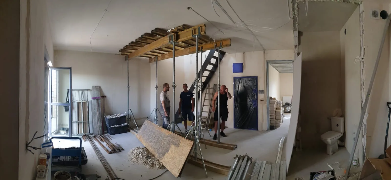 Panoramic view of duplex conversion showing ULMA timber shoring across slab opening, steel staircase being installed, three-person construction crew working between levels