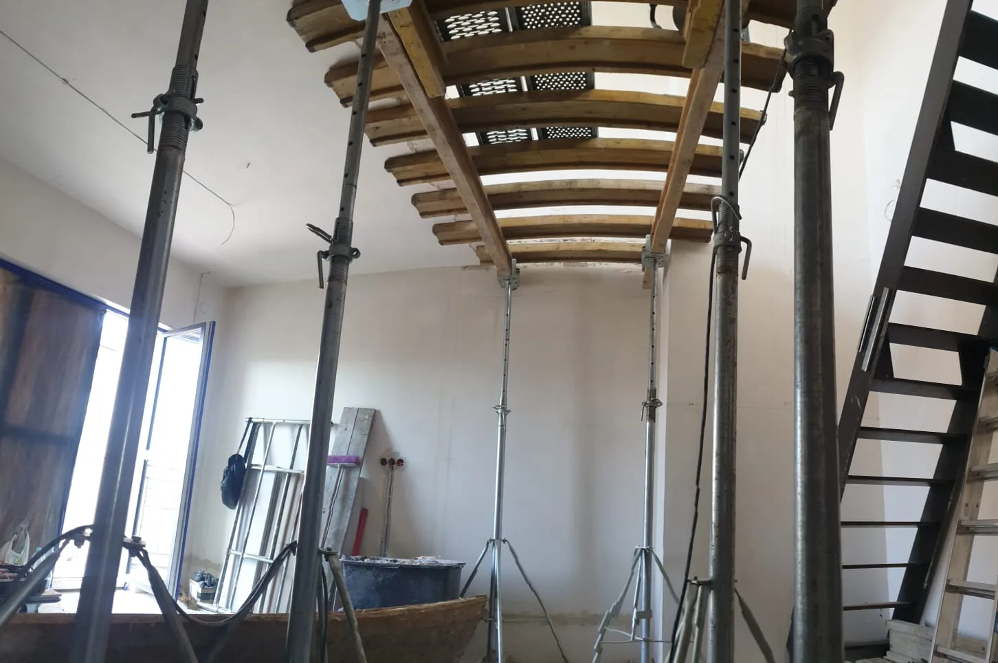 Ground-level view of ULMA shoring system with multiple acrow props supporting the cut slab opening, steel staircase visible beyond, construction equipment in foreground