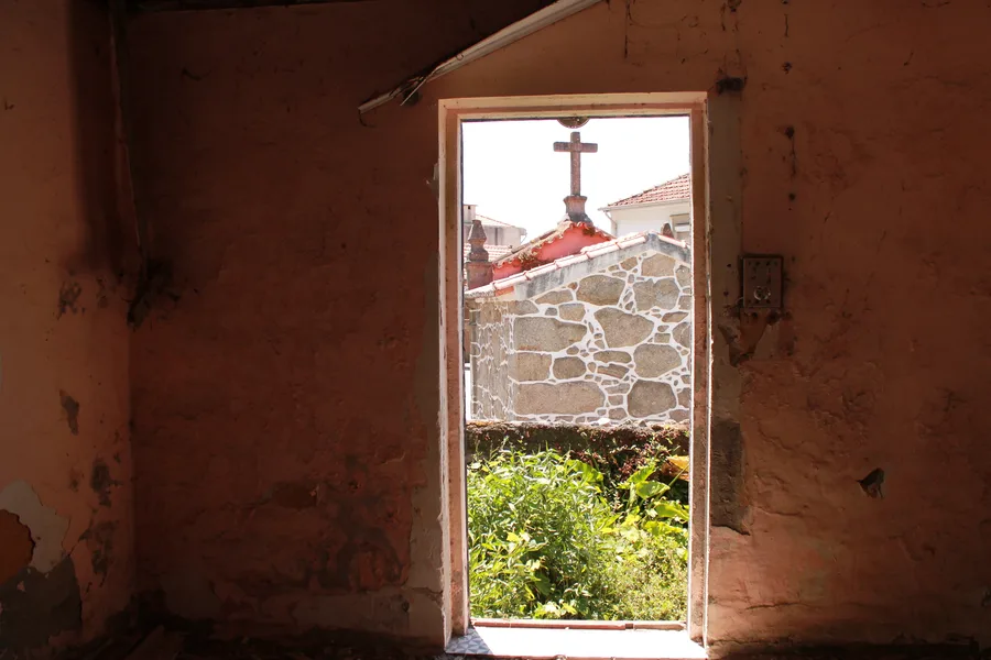 Before: interior photo looking out through granite masonry doorway with stone cross visible beyond, showing thick rough-hewn walls and collapsed timber roof structure above