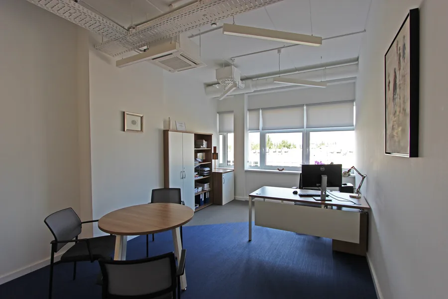Private office with round meeting table, desk at window, bookshelf and walnut wardrobe unit, framed certificates, and blue woven vinyl floor
