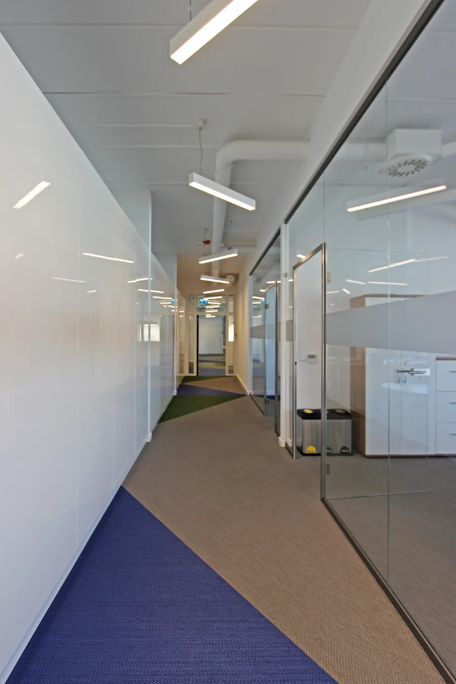 Portrait corridor view showing the rhythm of glass partitions and woven vinyl flooring extending into the depth of the office floor