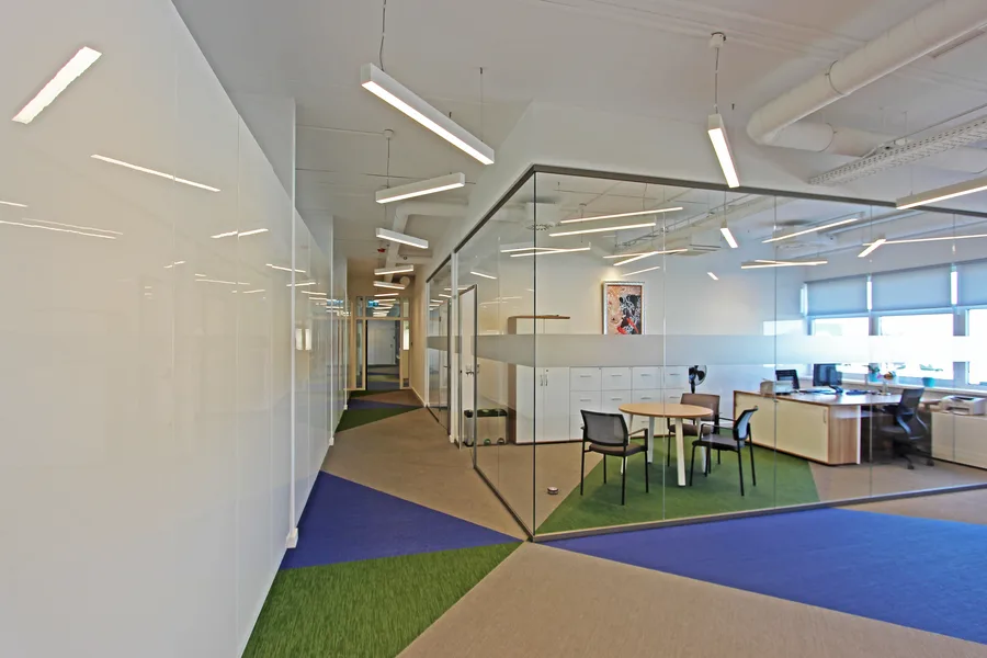 Corridor-to-office perspective with glass partition corner creating strong geometric composition, frosted privacy band, and woven vinyl floor colour-blocking