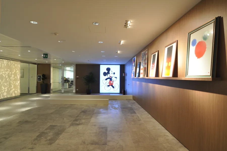 Art gallery corridor with Grey Oak veneer walls, framed Disney artwork on ledge shelving, and the dramatic backlit Mickey lightbox endpoint