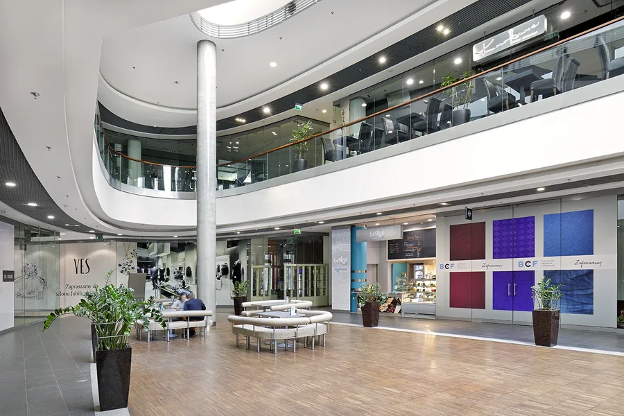 Interior: professional photograph of Drukarnia Mall common area showing ceiling system, retail unit entries, and wayfinding signage