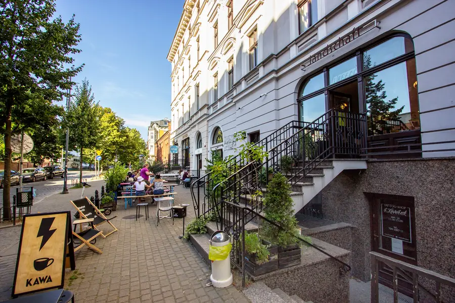 Summer street with KAWA coffee sign, deck chairs, restored facade, Landschaft entrance with iron staircase, potted plants, cobblestone