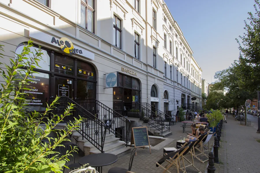 Summer street view with AleBra and BARZZA signage, outdoor seating with deck chairs, pedestrians relaxing, trees, multiple storefront entrances