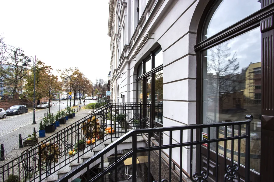 Autumn entrance stairs from elevated viewpoint with potted plants and pumpkins on steps, iron railings, large arched window, street with autumn trees