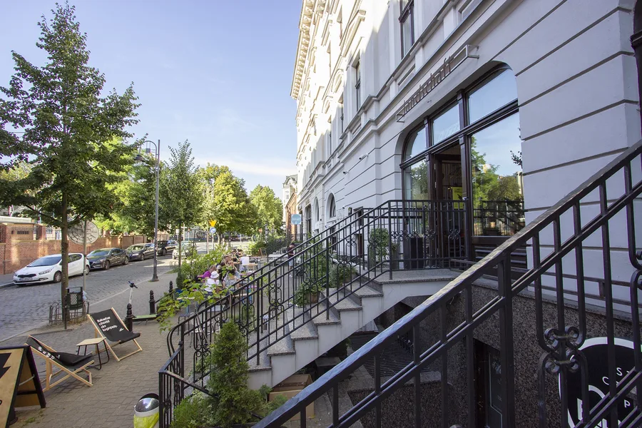 Entrance staircase from street level with white stone steps, iron railing, building corner with street view, cars and outdoor seating visible